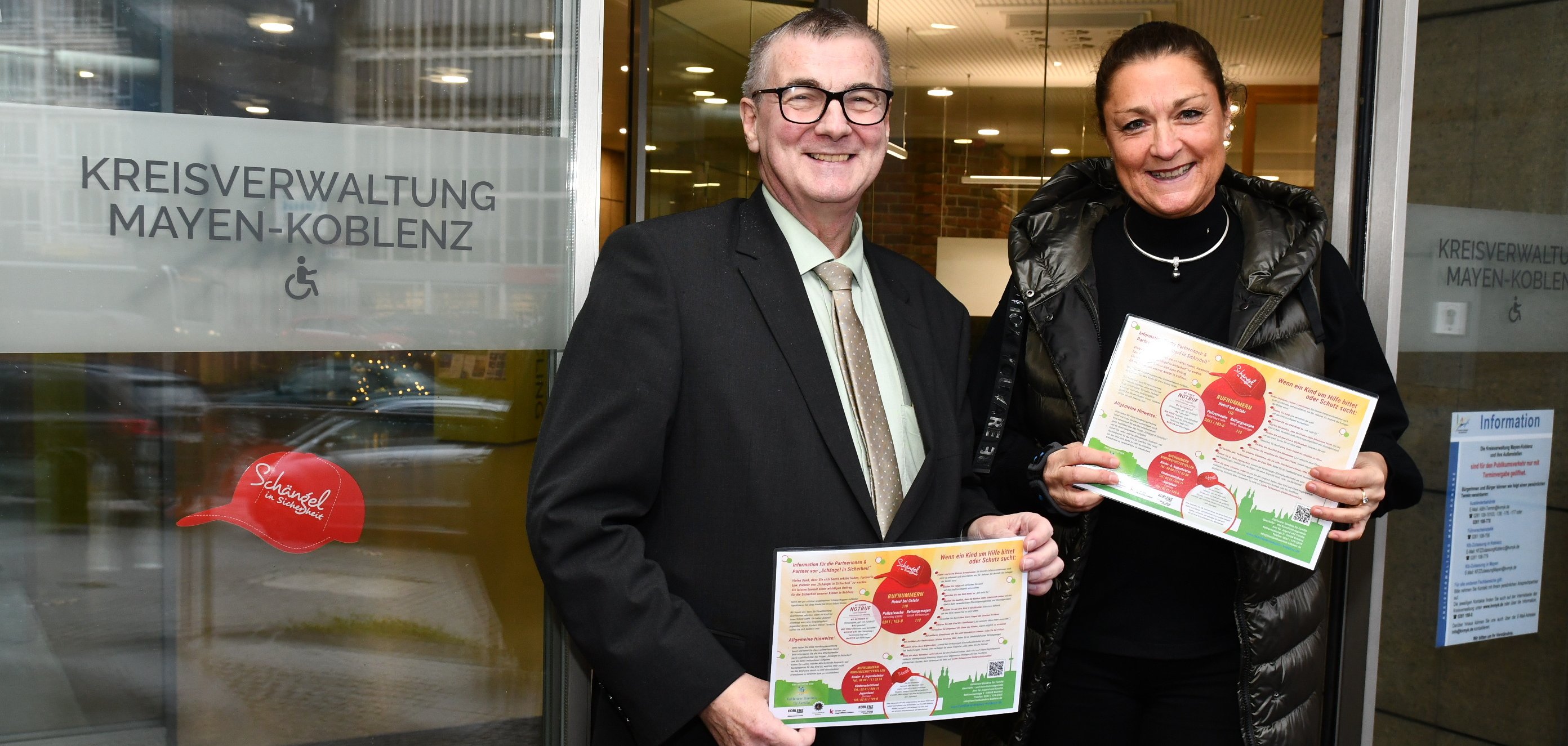 Landrat Dr. Alexander Saftig und Minka Bäumges vom Koblenzer Bündnis für Fami-lien haben gemeinsam einen Aufkleber in Form einer roten Kappe mit der Aufschrift „Schängel in Sicherheit“ am Eingang des Kreishauses angebracht. Die Kreisverwaltung ist somit fortan gut als sicherer Ort für Kinder in Notsituationen erkennbar.